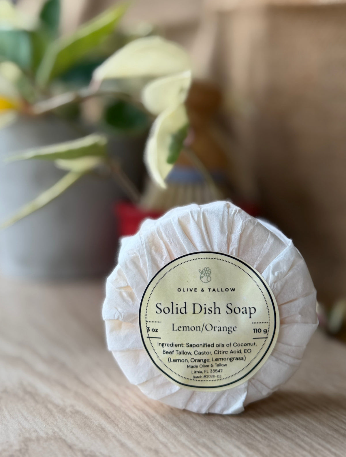 A wrapped bar of Olive & Tallow Solid Dish Soap in Lemon/Orange scent, displayed on a wooden surface with a blurred background of a plant and rustic wall.