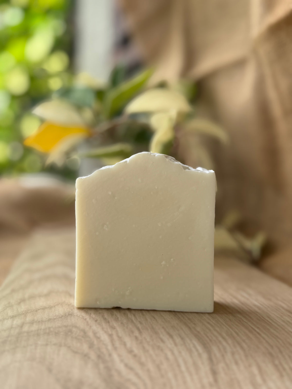 A rectangular bar of handmade soap with a rustic, slightly uneven top edge, standing upright on a light wooden surface. The soap has a matte, creamy off-white color.