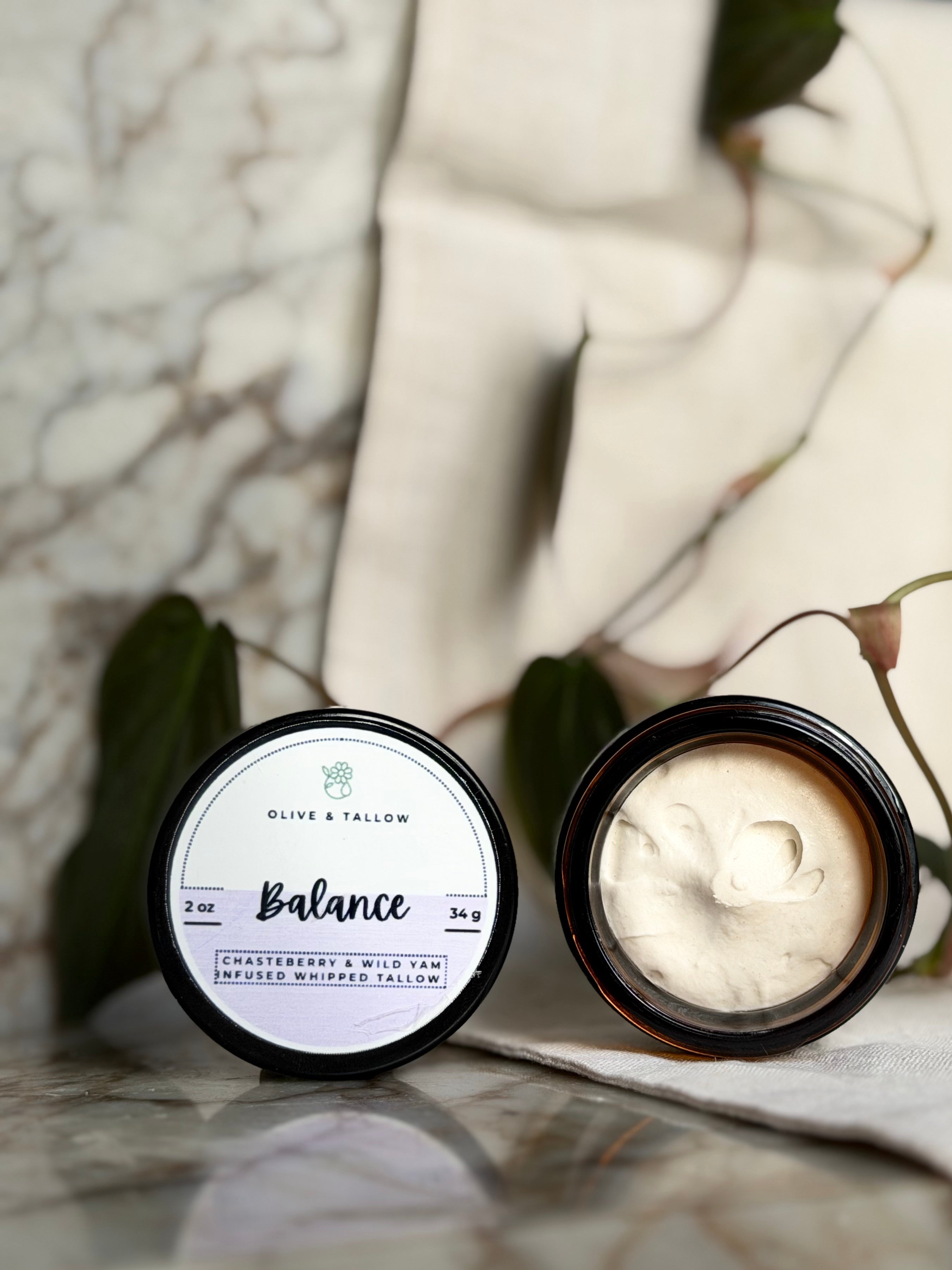 An open round jar of Olive & Tallow Balance balm, showing the creamy off-white product inside, with its lid placed beside it on a marble surface.