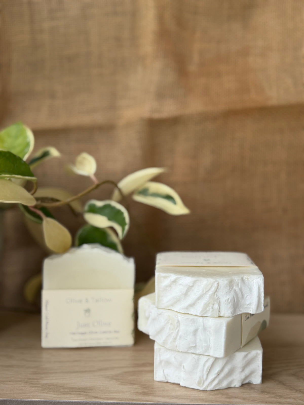 Three stacked white textured soap bars next to a single wrapped soap bar with a cream label, set on a wooden surface with a blurred burlap backdrop and variegated plant leaves in the background.